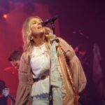 Cantora Anne Wilson se apresenta no palco, loira, de blusa branca, diante do microfone sob luzes vermelhas.