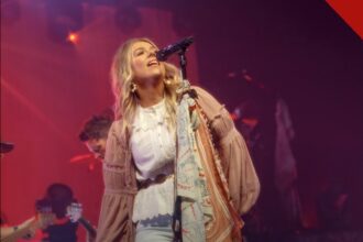 Cantora Anne Wilson se apresenta no palco, loira, de blusa branca, diante do microfone sob luzes vermelhas.