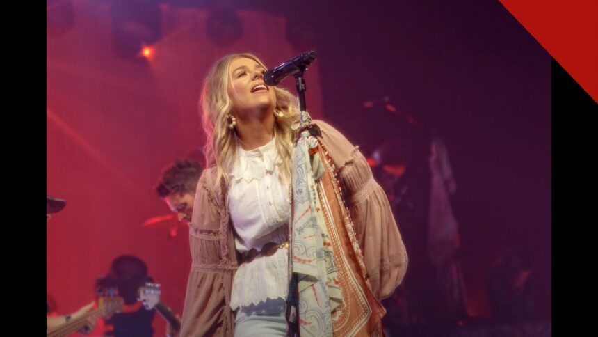 Cantora Anne Wilson se apresenta no palco, loira, de blusa branca, diante do microfone sob luzes vermelhas.