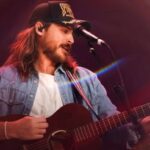 Cory Asbury de boné e camisa jeans tocando violão e cantando no palco durante um show