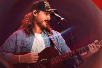 Cory Asbury de boné e camisa jeans tocando violão e cantando no palco durante um show