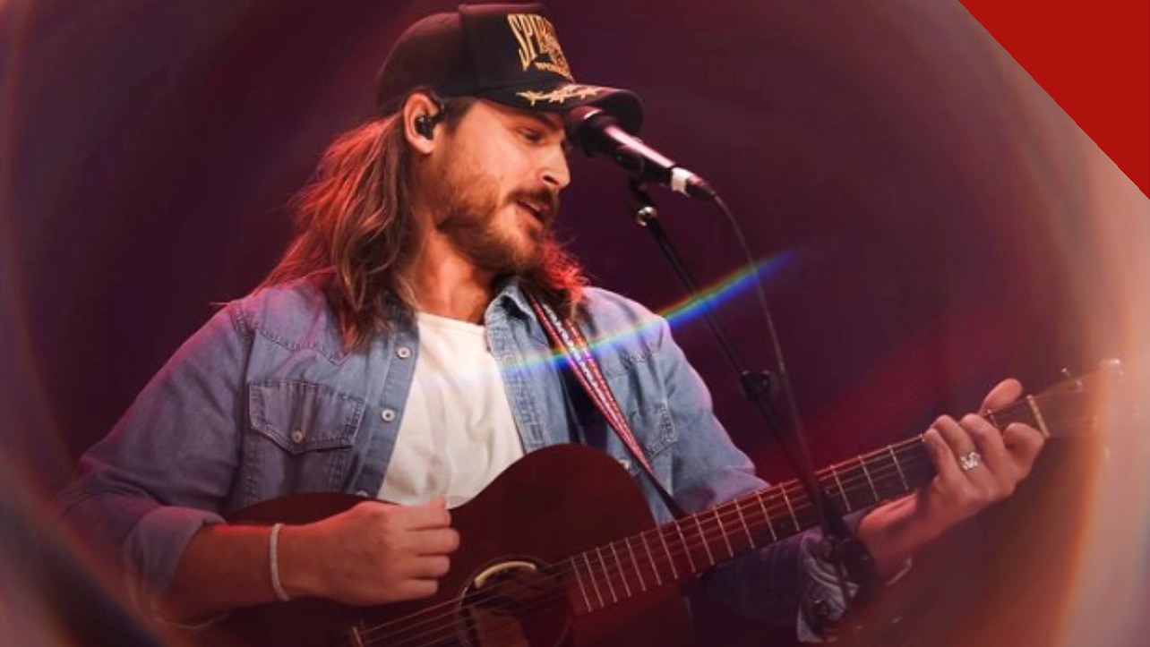 Cory Asbury de boné e camisa jeans tocando violão e cantando no palco durante um show