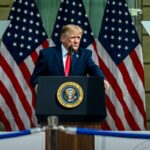 Donald Trump discursa em púlpito presidencial diante de três bandeiras dos Estados Unidos.