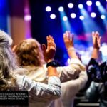 Fiéis de igreja com as mãos levantadas em adoração sob luzes coloridas durante um momento de oração