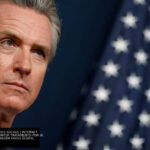 Governador Gavin Newsom, da Califórnia, em evento oficial com a bandeira dos Estados Unidos ao fundo.