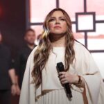 A cantora gospel Sarah Farias se apresenta de vestido branco com microfone, sob luzes rosas no palco