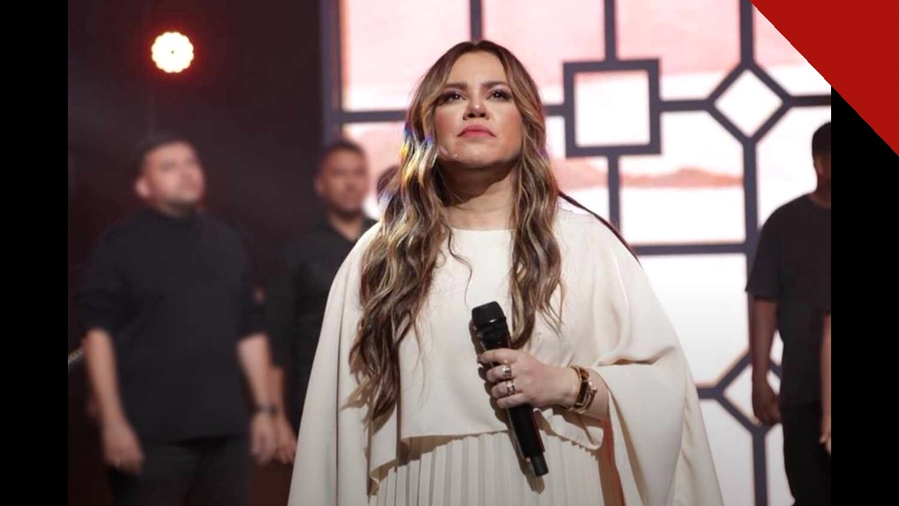 A cantora gospel Sarah Farias se apresenta de vestido branco com microfone, sob luzes rosas no palco