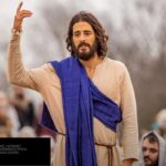 Jonathan Roumie caracterizado como Jesus em cena de The Chosen, com túnica e manto azul, pregando para uma multidão