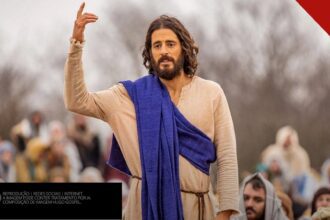 Jonathan Roumie caracterizado como Jesus em cena de The Chosen, com túnica e manto azul, pregando para uma multidão
