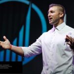 Tim Tebow de camisa branca palestrando no palco da Life Surge, com a marca da empresa ao fundo
