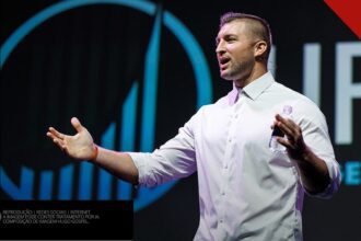 Tim Tebow de camisa branca palestrando no palco da Life Surge, com a marca da empresa ao fundo