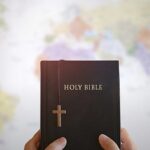 igrejas do Reino Unido gastam menos com evangelismo: mãos segurando uma Bíblia com uma cruz de madeira diante de um mapa do mundo
