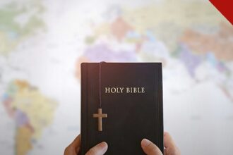 igrejas do Reino Unido gastam menos com evangelismo: mãos segurando uma Bíblia com uma cruz de madeira diante de um mapa do mundo