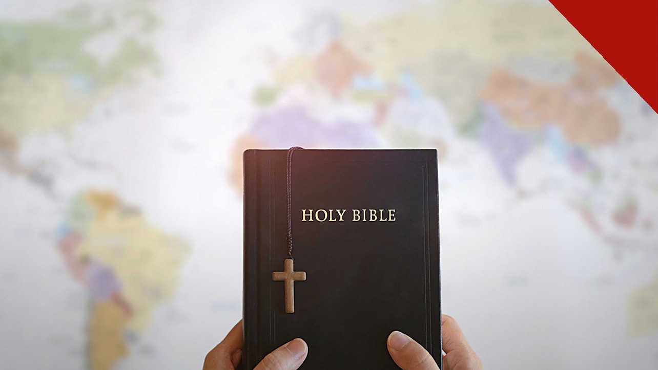 igrejas do Reino Unido gastam menos com evangelismo: mãos segurando uma Bíblia com uma cruz de madeira diante de um mapa do mundo