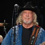 John Anderson morre aos 71 anos: cantor country de chapéu preto e guitarra no palco durante show
