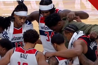 Jogadores do Washington Wizards de cabeça baixa em círculo de oração com Barnes no meio da quadra, em imagem que ilustra como jogadores do Wizards oram com Scottie Barnes após a partida
