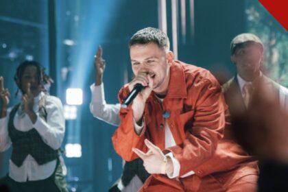 Hulvey cancela show: o rapper cristão Hulvey canta com microfone em apresentação, vestindo roupa vermelha, com dançarinos ao fundo