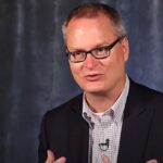 igreja de Adam Hamilton usa termos neutros: o pastor Adam Hamilton, de óculos e blazer escuro, falando em entrevista com fundo azul