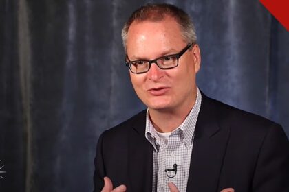 igreja de Adam Hamilton usa termos neutros: o pastor Adam Hamilton, de óculos e blazer escuro, falando em entrevista com fundo azul