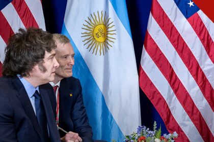 Milei vai à inauguração de igreja evangélica: presidente argentino de perfil, com as bandeiras da Argentina e dos Estados Unidos ao fundo