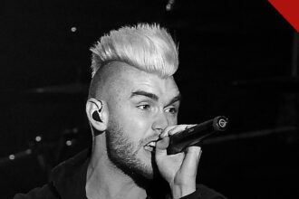 Colton Dixon fala da música Bones: cantor de moicano claro cantando ao microfone em show, em preto e branco