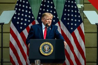 Conselho de Paz de Trump nomeia ex oficial de Biden é a decisão do Conselho, e a imagem mostra o presidente Donald Trump falando em púlpito oficial com bandeiras dos Estados Unidos