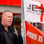 Igreja britânica contesta proibição de pregação: pregador com microfone diante de faixas com o nome de Jesus em rua de Colchester