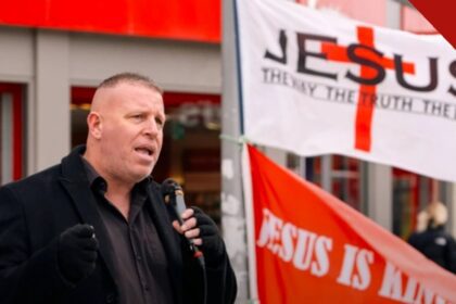 Igreja britânica contesta proibição de pregação: pregador com microfone diante de faixas com o nome de Jesus em rua de Colchester
