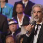 Guy Penrod enfrenta diagnóstico grave de saúde, e a imagem mostra o cantor Guy Penrod, de cabelos longos grisalhos, cantando com um microfone na mão diante de um coral
