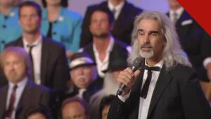 Guy Penrod enfrenta diagnóstico grave de saúde, e a imagem mostra o cantor Guy Penrod, de cabelos longos grisalhos, cantando com um microfone na mão diante de um coral