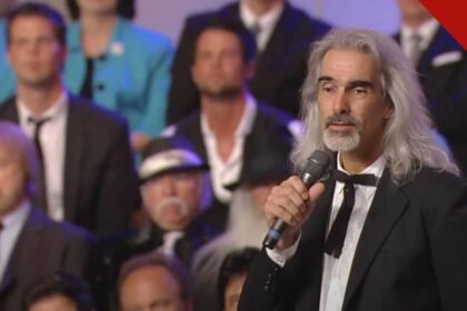 Guy Penrod enfrenta diagnóstico grave de saúde, e a imagem mostra o cantor Guy Penrod, de cabelos longos grisalhos, cantando com um microfone na mão diante de um coral