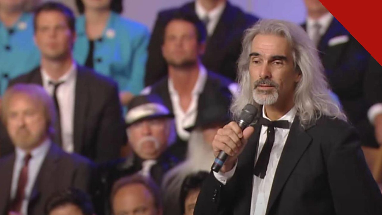 Guy Penrod enfrenta diagnóstico grave de saúde, e a imagem mostra o cantor Guy Penrod, de cabelos longos grisalhos, cantando com um microfone na mão diante de um coral