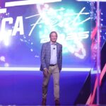 Pastores apoiam Ken Paxton no Texas: Ken Paxton sorrindo de corpo inteiro no palco de evento público, com painel luminoso ao fundo