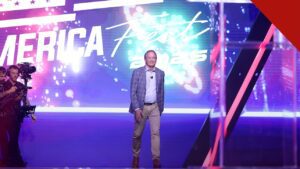 Pastores apoiam Ken Paxton no Texas: Ken Paxton sorrindo de corpo inteiro no palco de evento público, com painel luminoso ao fundo