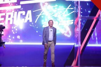 Pastores apoiam Ken Paxton no Texas: Ken Paxton sorrindo de corpo inteiro no palco de evento público, com painel luminoso ao fundo