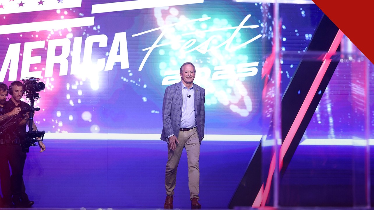 Pastores apoiam Ken Paxton no Texas: Ken Paxton sorrindo de corpo inteiro no palco de evento público, com painel luminoso ao fundo