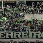 Jakson Follmann relembra como soube das mortes dos companheiros, e a imagem mostra a torcida da Chapecoense com cachecóis e a faixa Guerreiros em tributo na Arena Condá