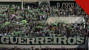 Jakson Follmann relembra como soube das mortes dos companheiros, e a imagem mostra a torcida da Chapecoense com cachecóis e a faixa Guerreiros em tributo na Arena Condá