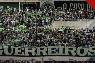 Jakson Follmann relembra como soube das mortes dos companheiros, e a imagem mostra a torcida da Chapecoense com cachecóis e a faixa Guerreiros em tributo na Arena Condá
