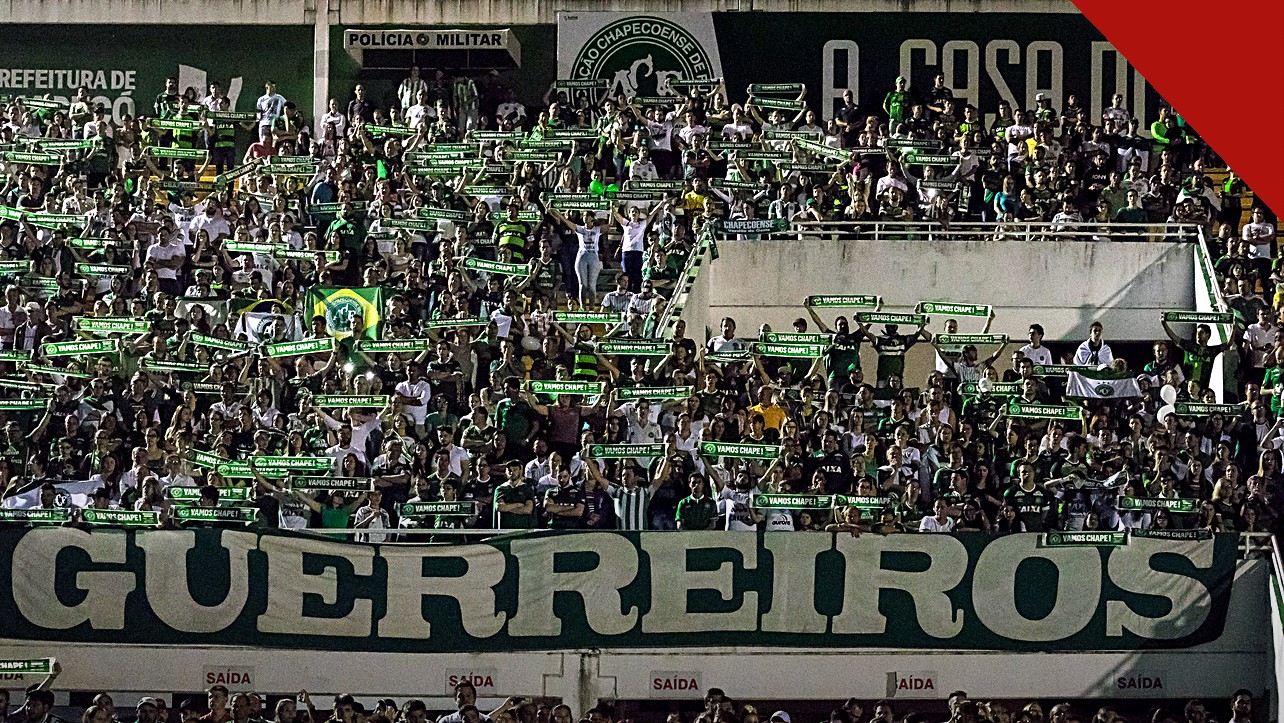 Jakson Follmann relembra como soube das mortes dos companheiros, e a imagem mostra a torcida da Chapecoense com cachecóis e a faixa Guerreiros em tributo na Arena Condá