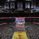 Ernie Johnson deixa o Inside the NBA após 3 décadas, e a imagem mostra o interior de uma arena da NBA lotada, com a quadra de basquete ao centro e faixas de campeões no teto