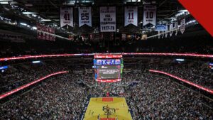Ernie Johnson deixa o Inside the NBA após 3 décadas, e a imagem mostra o interior de uma arena da NBA lotada, com a quadra de basquete ao centro e faixas de campeões no teto