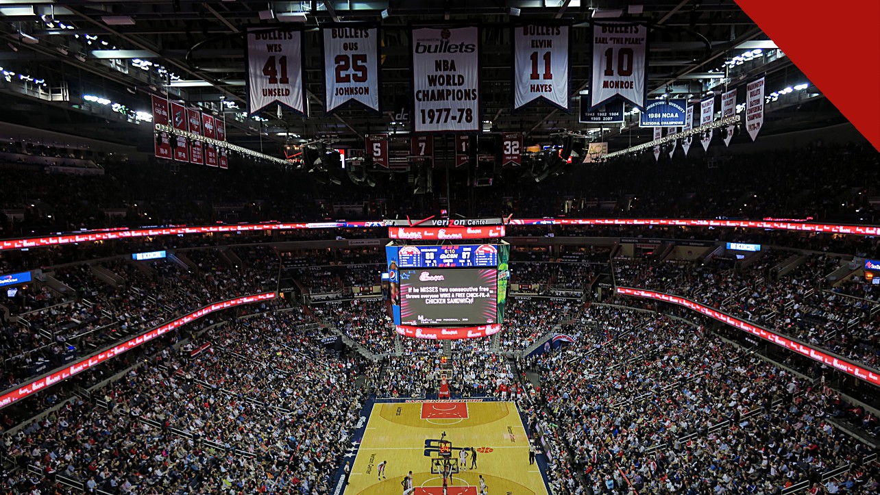 Ernie Johnson deixa o Inside the NBA após 3 décadas, e a imagem mostra o interior de uma arena da NBA lotada, com a quadra de basquete ao centro e faixas de campeões no teto