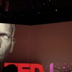 polícia desarticula esquema com deepfake de famosos, e a imagem mostra uma demonstração de deepfake em telão no TED 2019, com um rosto digital projetado ao lado do apresentador