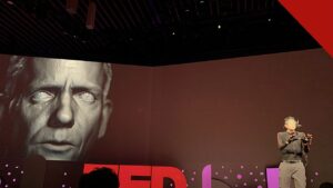 polícia desarticula esquema com deepfake de famosos, e a imagem mostra uma demonstração de deepfake em telão no TED 2019, com um rosto digital projetado ao lado do apresentador