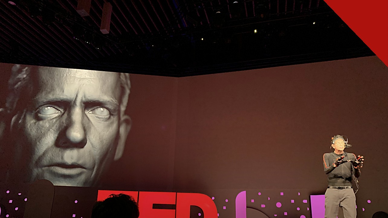polícia desarticula esquema com deepfake de famosos, e a imagem mostra uma demonstração de deepfake em telão no TED 2019, com um rosto digital projetado ao lado do apresentador