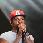 Kirk Franklin lança álbum duplo como resposta aos críticos, e a imagem mostra Kirk Franklin cantando em um palco, de boné vermelho e camiseta branca, segurando um microfone com as duas mãos