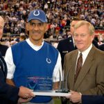 Tony Dungy repreende pastor que defende poligamia, e a imagem mostra Tony Dungy, de colete azul do Indianapolis Colts, recebendo uma homenagem em campo, cercado por oficiais e convidados