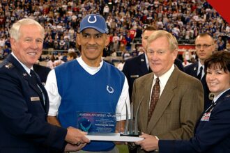Tony Dungy repreende pastor que defende poligamia, e a imagem mostra Tony Dungy, de colete azul do Indianapolis Colts, recebendo uma homenagem em campo, cercado por oficiais e convidados