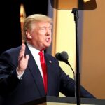políticas de Trump reduzem crescimento da população migrante, e a imagem mostra Donald Trump discursando em um púlpito, de terno escuro e gravata vermelha, com a mão levantada e bandeiras dos Estados Unidos ao fundo
